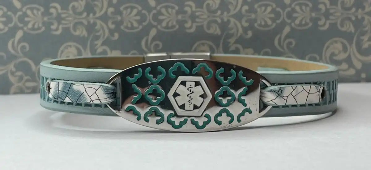 A light blue leather medical alert bracelet with a silver oval plate featuring a teal-colored medical symbol and decorative cutouts.